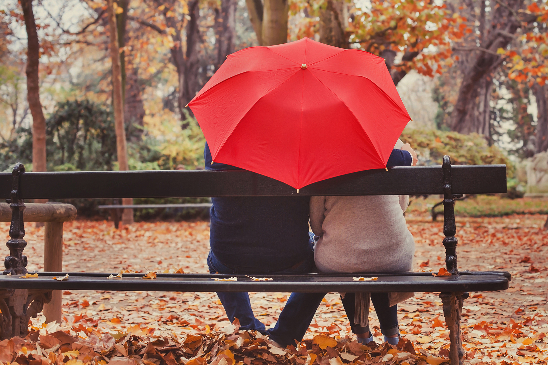 A senior couple sit on a park bench in fall beneath a red umbrella