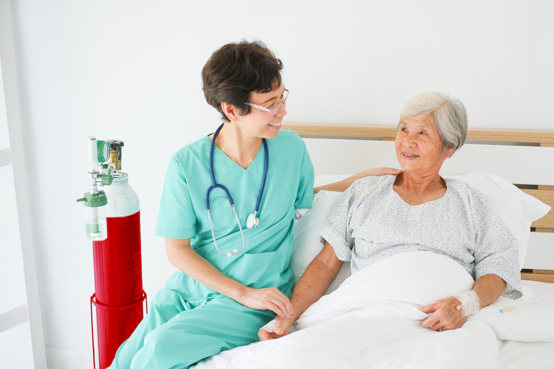 An Asian nurse in blue scrubs visits with an elderly female Asian patient, with a red oxygen tank nearby.