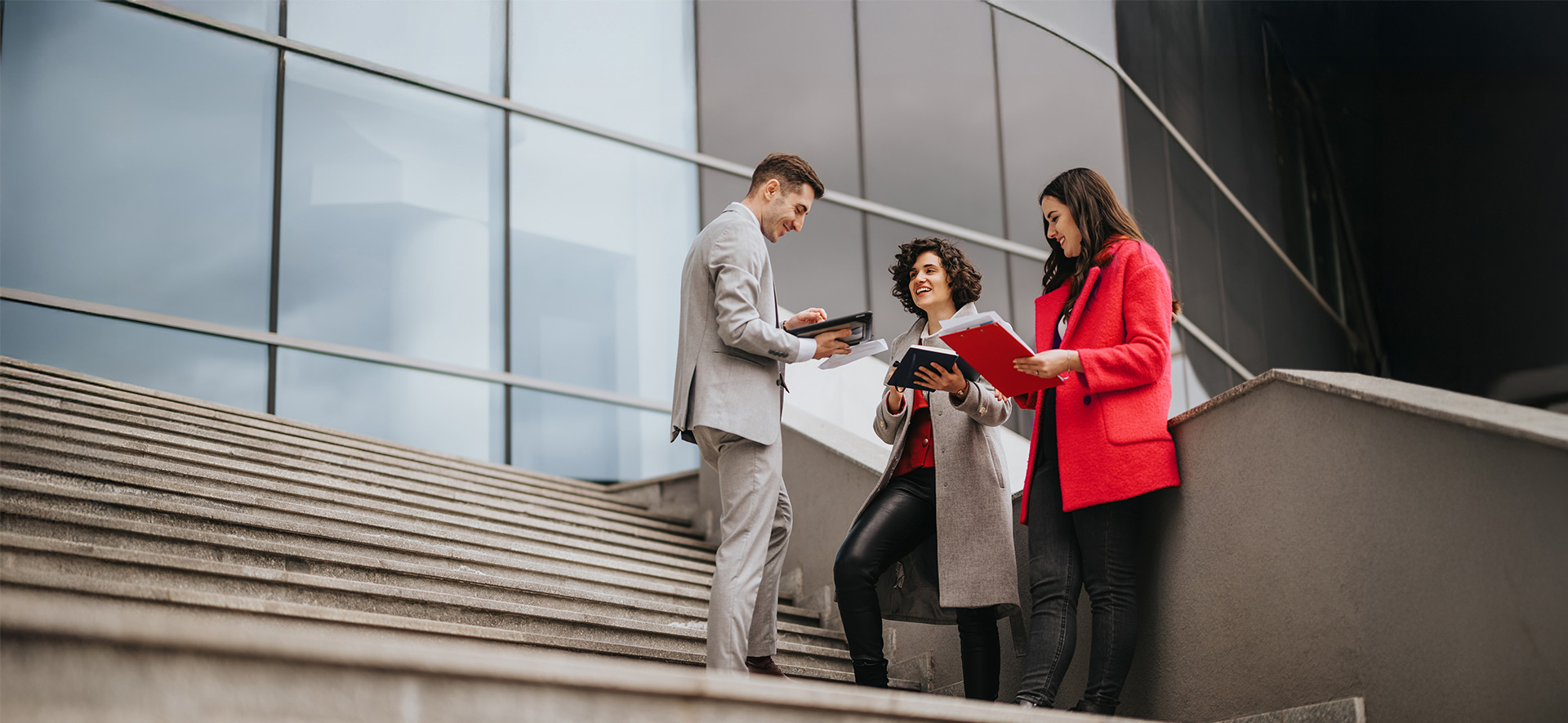 A group of three professionals standing on an outdoor staircase near a modern glass building, reviewing documents and discussing work