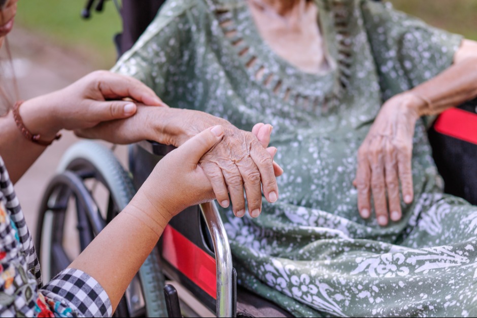 An elderly woman rests on a wheelchair, with a caregiver providing assistance