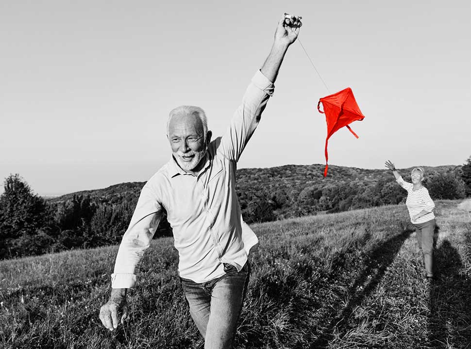 A smiling older man runs through a grassy hillside field flying a red kite while an older woman reaches up toward it, in a black-and-white photo with the kite highlighted in red.