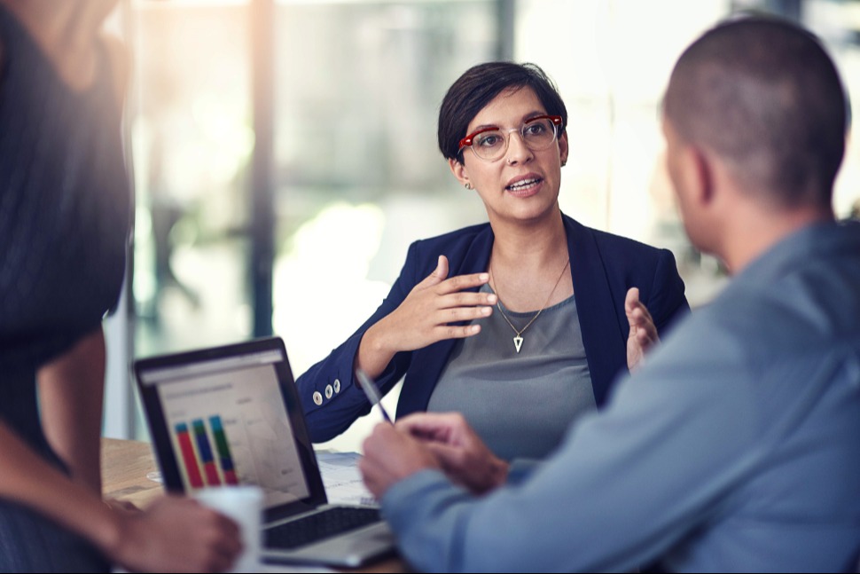 A specialist leans into a working conversation, red-framed glasses catching the light — the human read behind a market-entry decision
