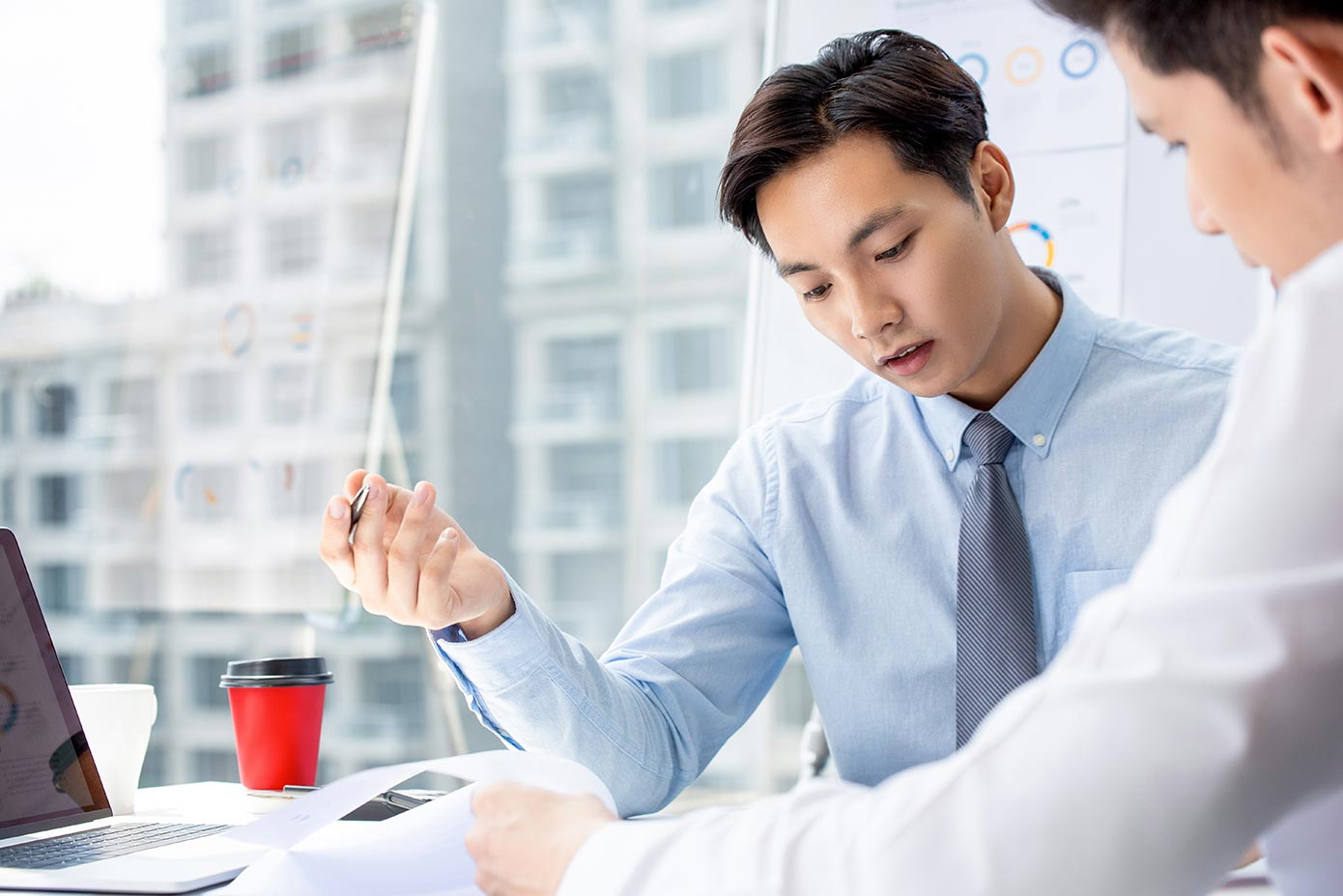 Two colleagues work through detail at a desk above a city skyline, a single red cup marking the table — the close analysis behind a cross-market longevity transaction.