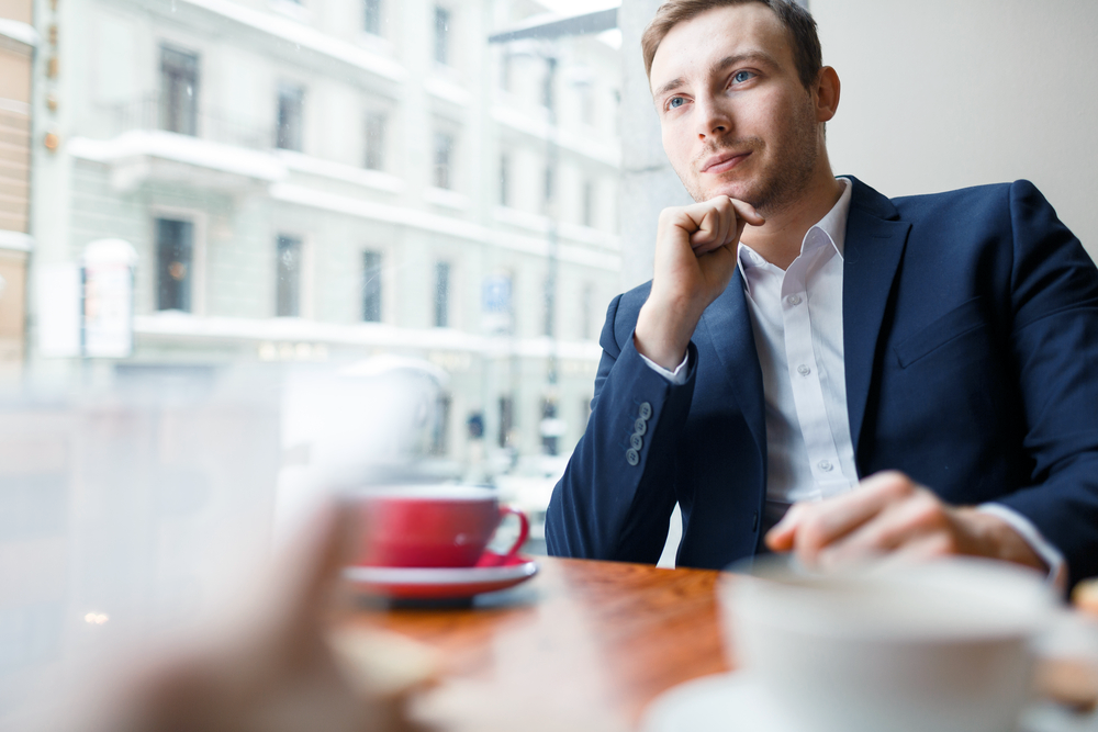 A figure pauses in thought by a bright window, a single red cup resting in the foreground — the considered thinking behind a pioneering capital structure.