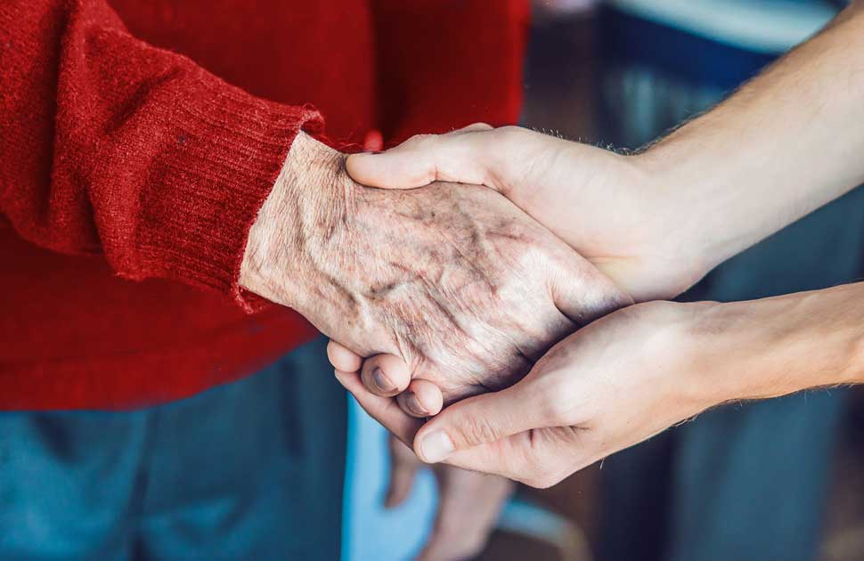 A young caregiver clutches the hand of an elderly woman wearing a red sweater