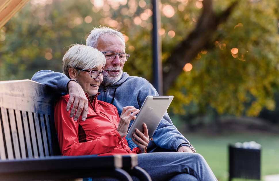 A retired couple sit close together on a park bench at golden hour, leaning over a tablet — the security of a future already settled.