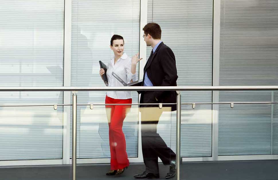 A pair of professionals stroll down a bridge, with a woman in red pants gesturing toward a man in a suit