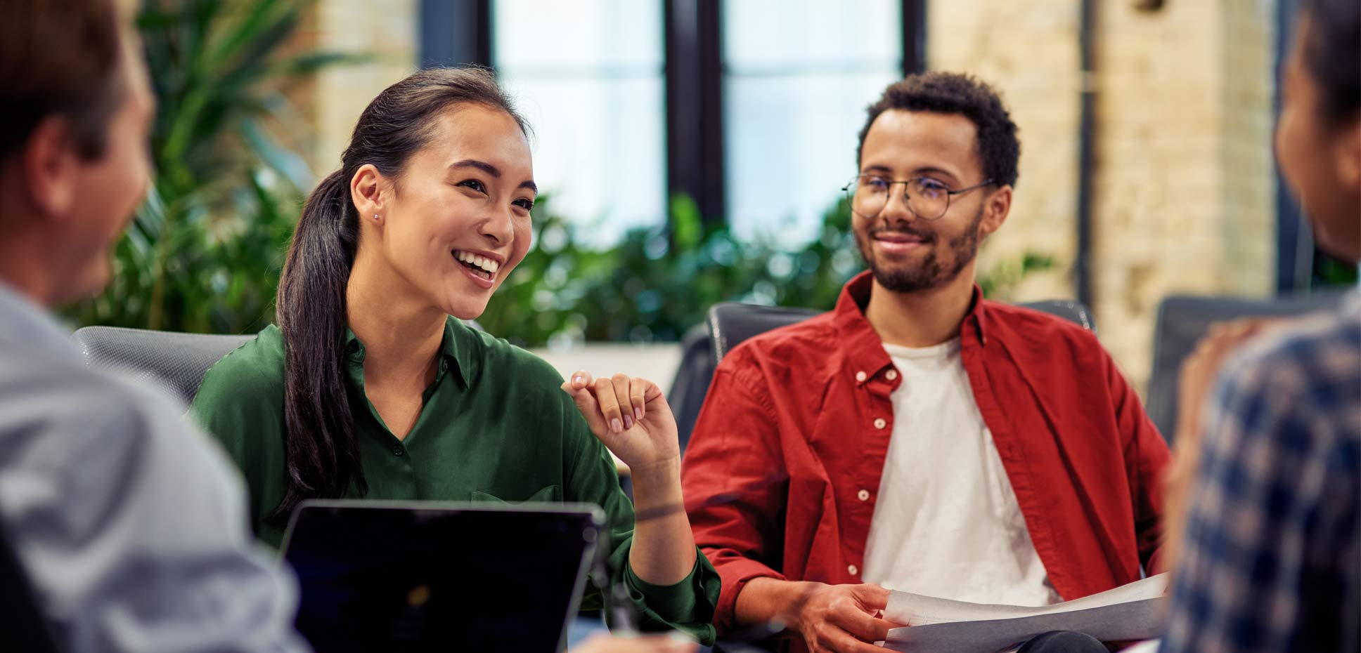 A female professional with a computer in her lap laughs as she speaks to a group of her peers