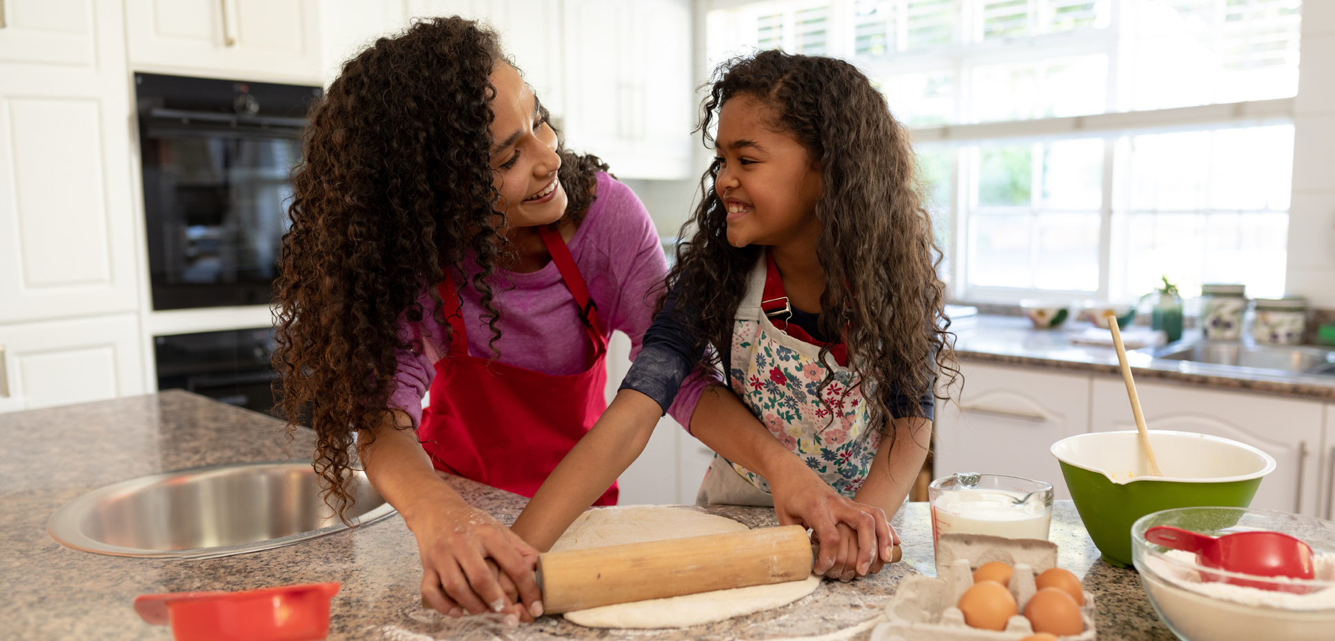 A smiling mother and young daughter roll out cookie dough together with a rolling pin in a sunny kitchen