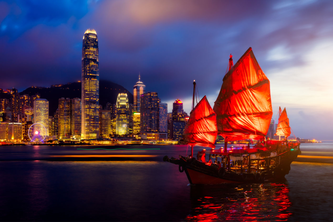 A traditional junk boat with red sails cruises the Hong Kong harbor at sunset