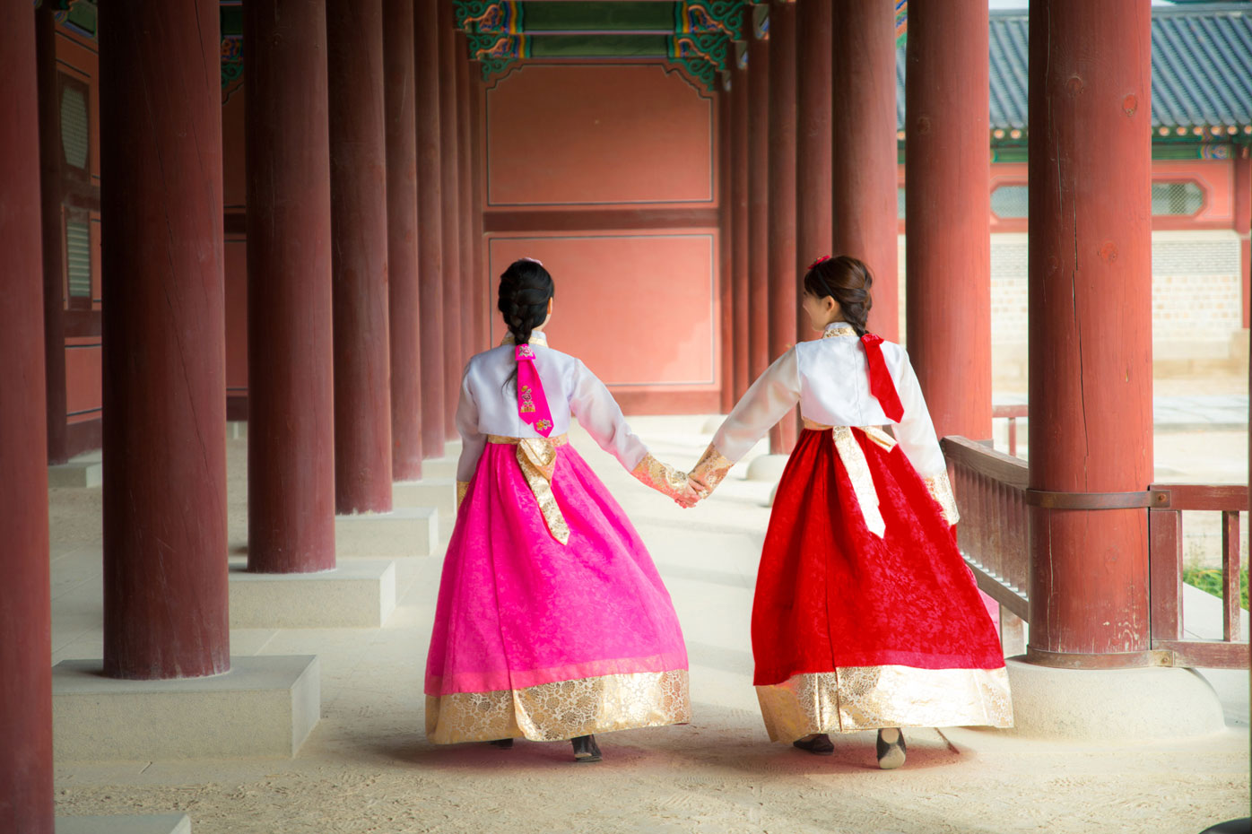 Two Korean women in traditional dresses hold hands while walking through a palace or temple