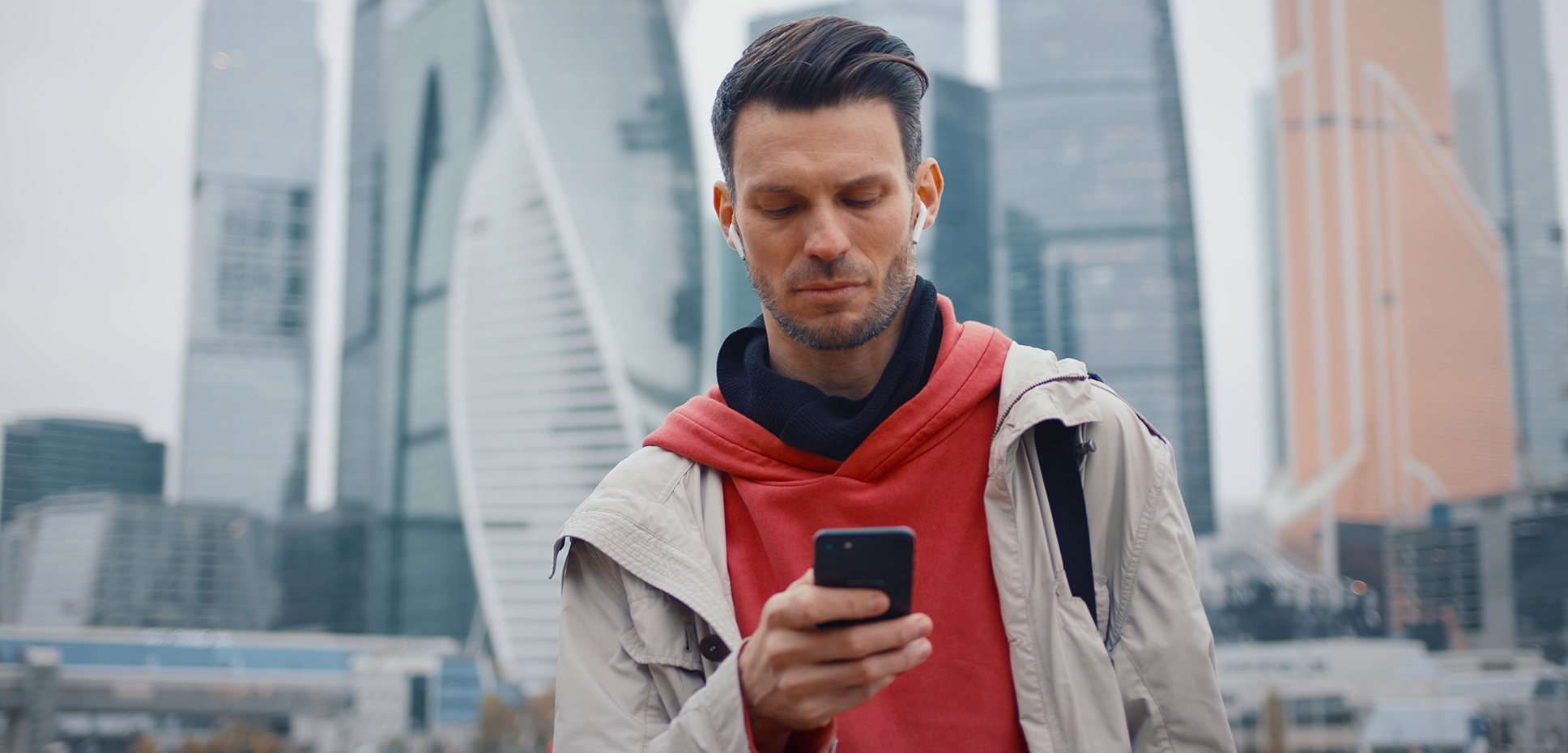 An urban commuter in a red hoodie and earbuds glances at his smartphone