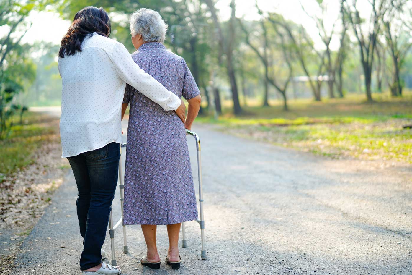 A caregiver assists an elderly woman enjoy a park while moving with a walker