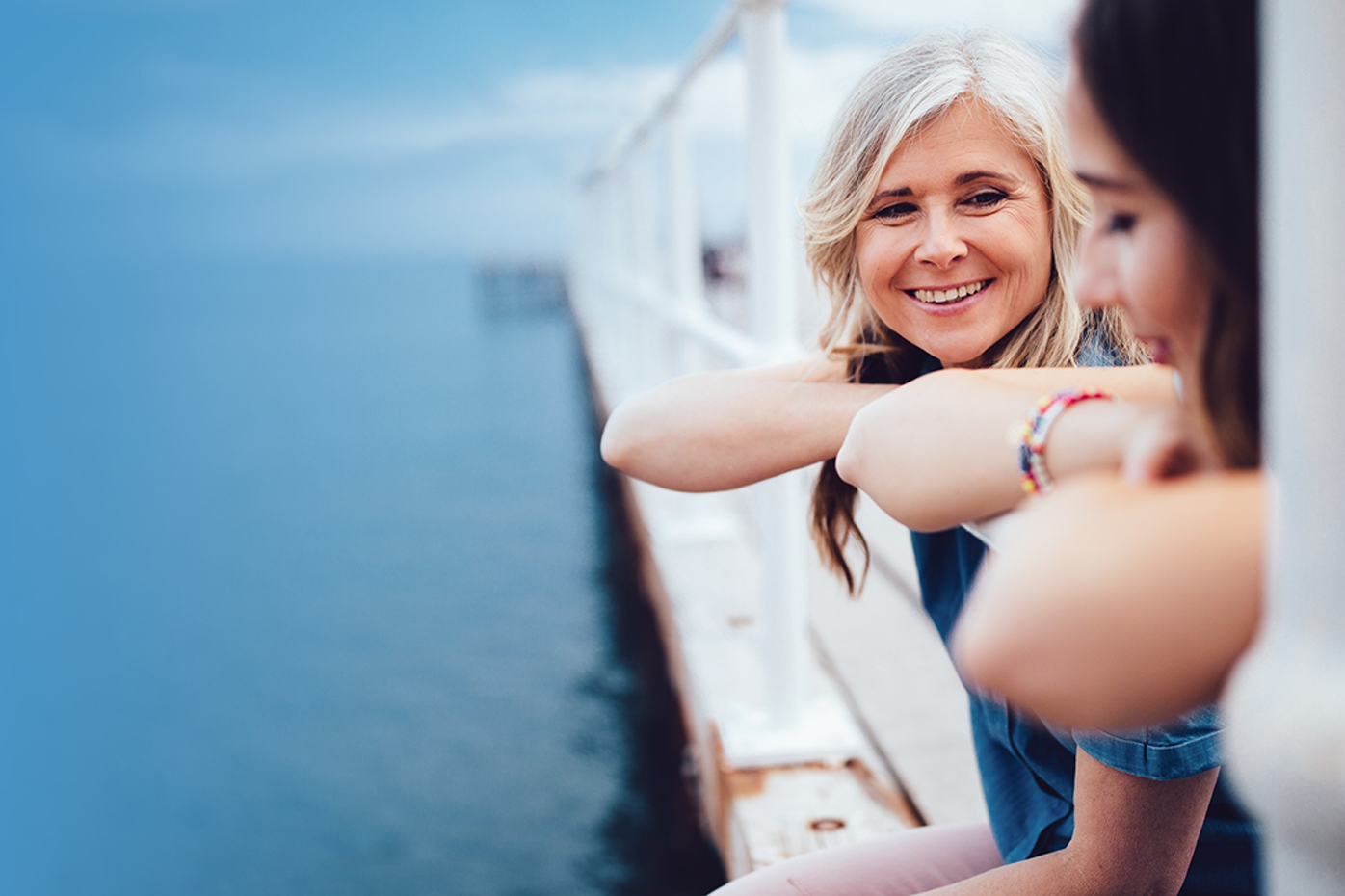 A pair of women lean over a pier, gazing at the water