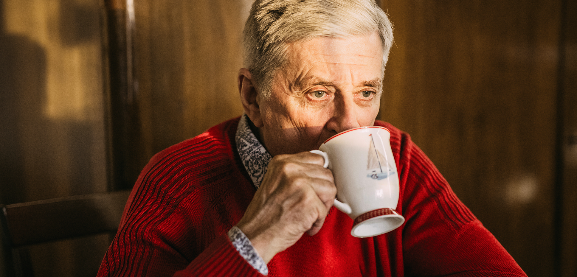 An older man wearing a bright red sweater pensively sips a cup of coffee.