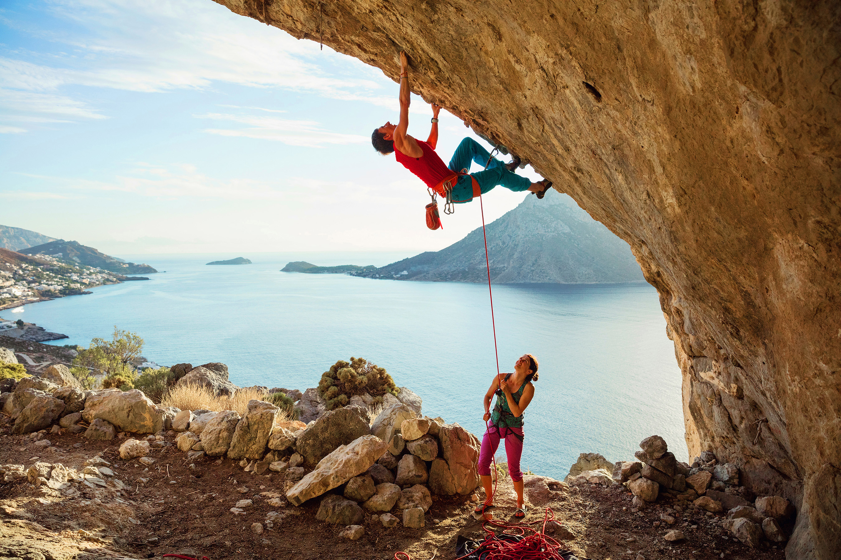 Man climbing cliff while woman holds safety rope below