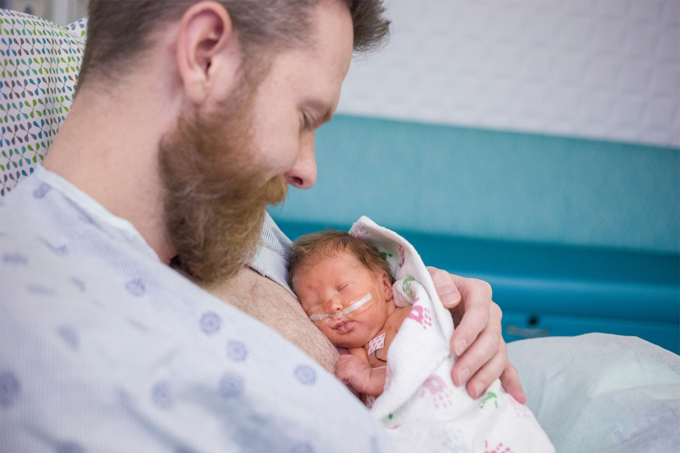 A father holds a preterm baby next to his bare chest in a hospital