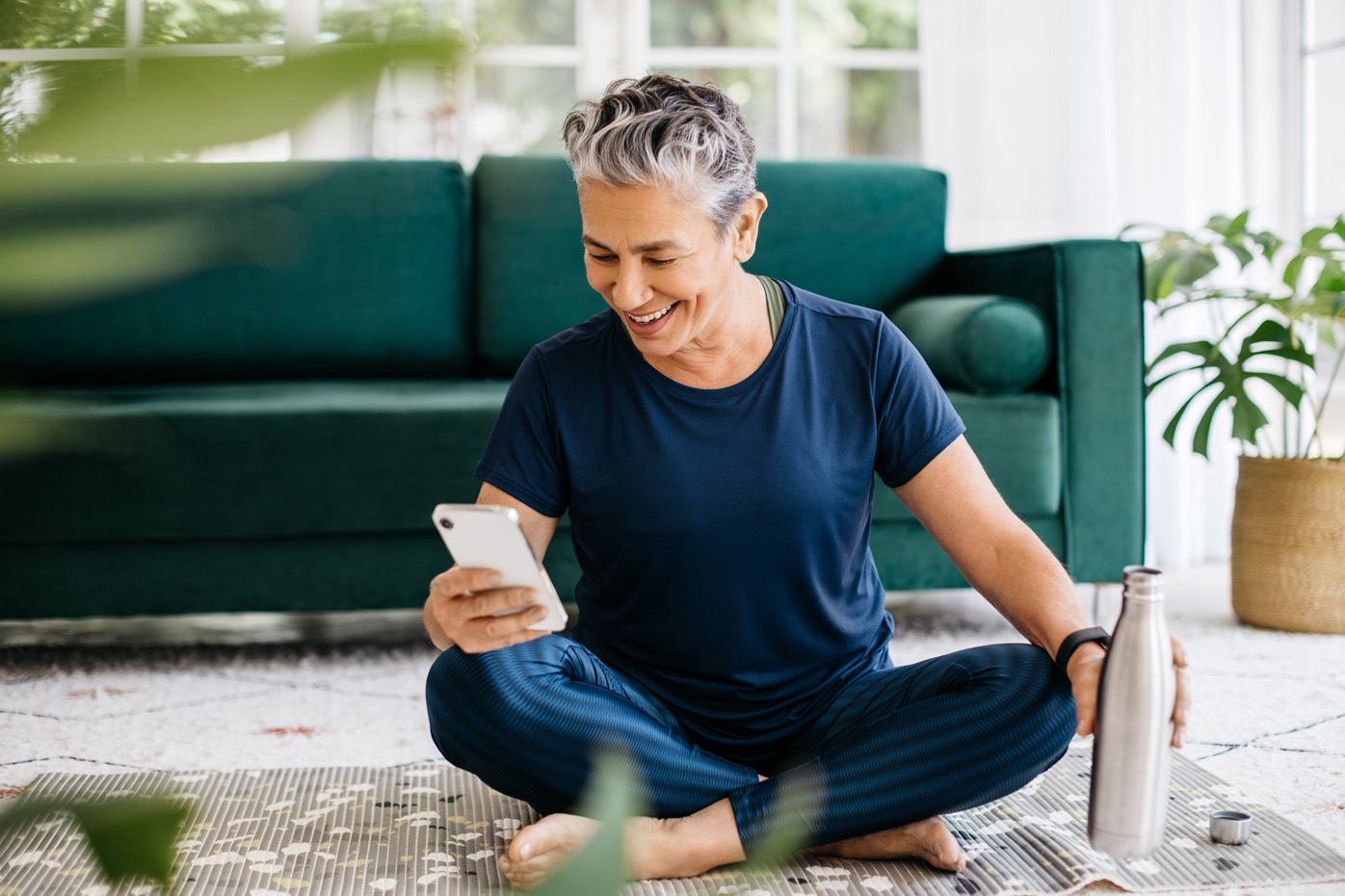 An older woman smiles and looks at a health app on her phone while sitting on a yoga mat and holding a silver water bottle.