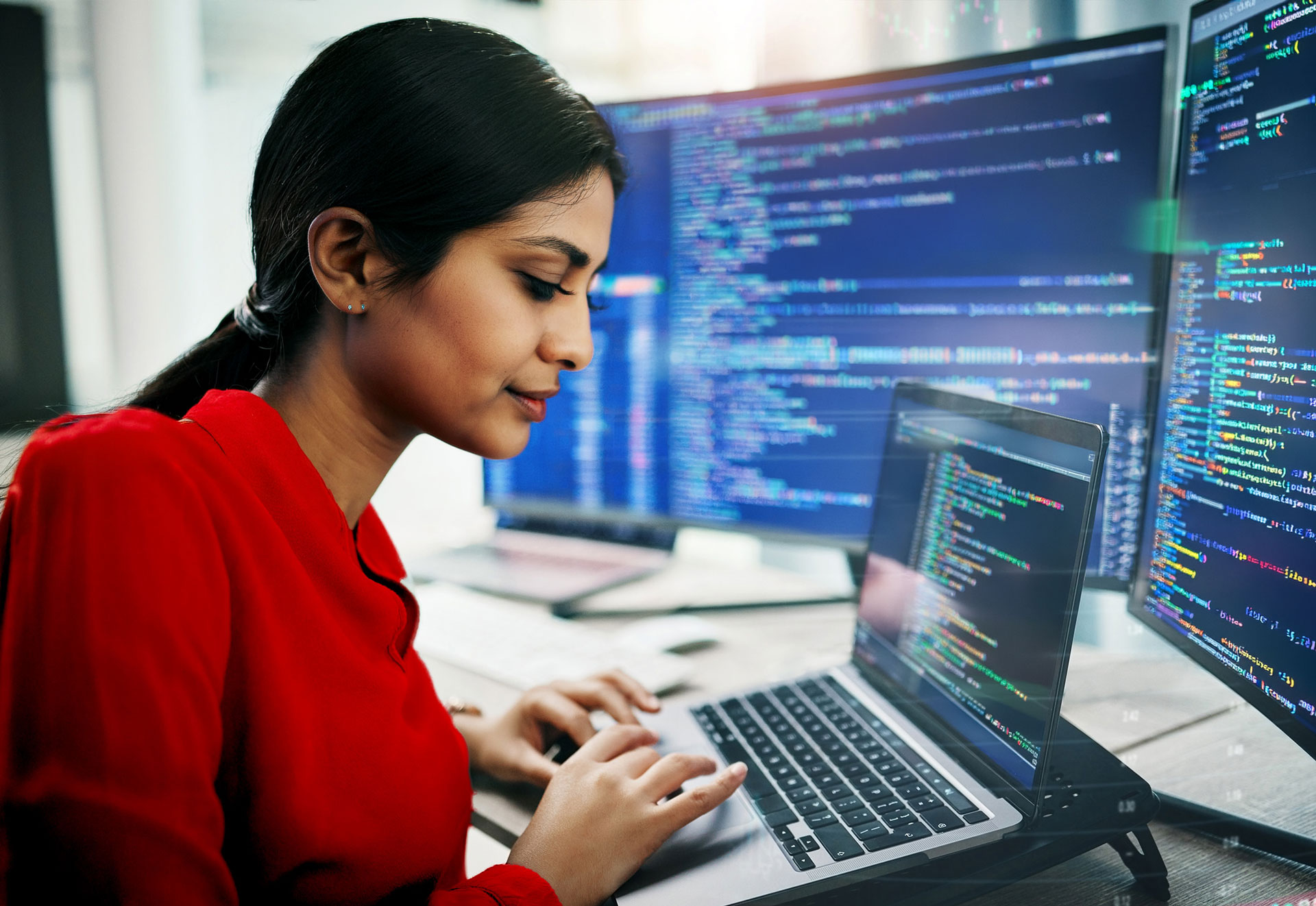 Person working on a laptop with multiple monitors displaying lines of code in a modern office setting.