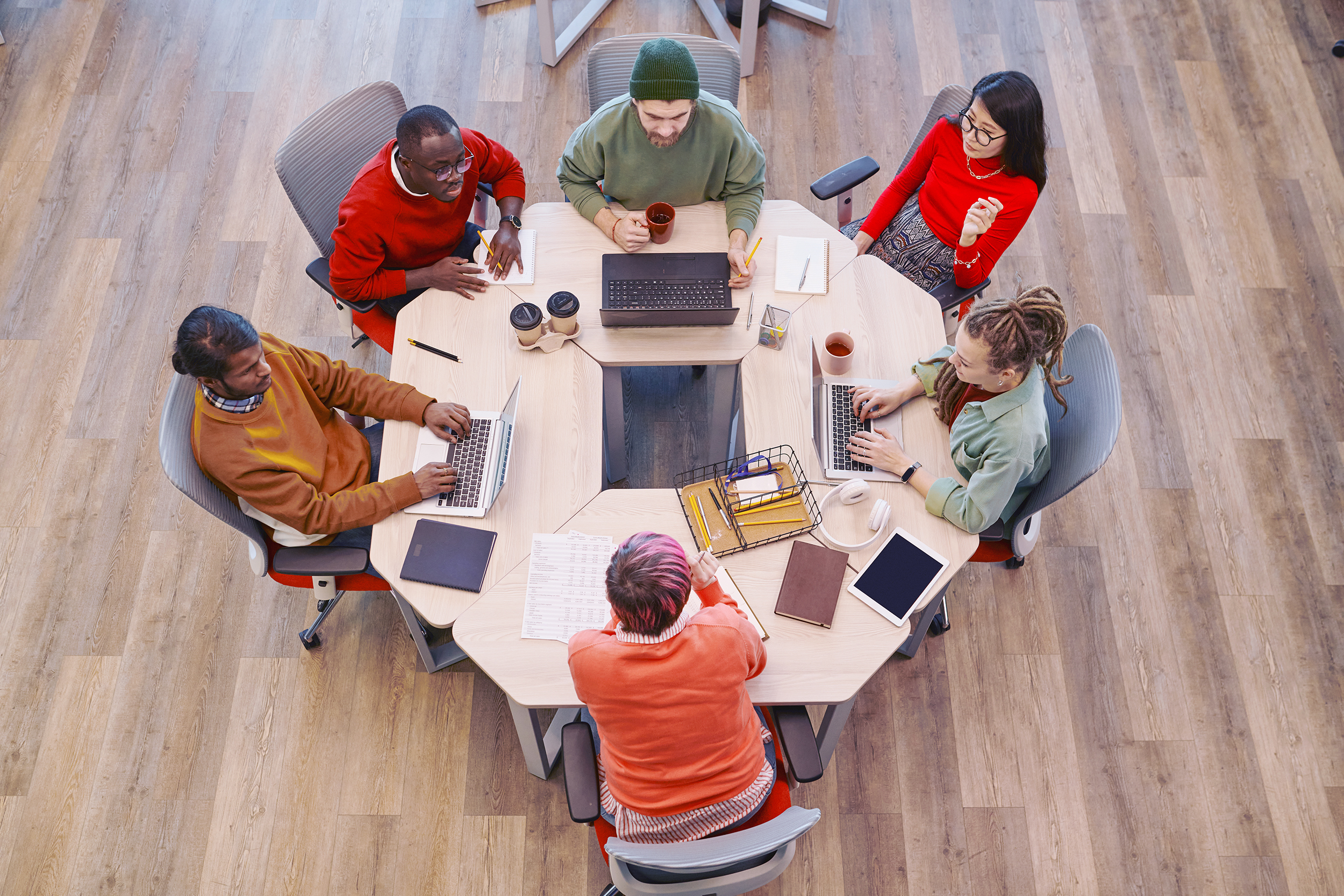 Six people seated around a modern hexagonal table, working on laptops and notebooks with coffee cups nearby, in a bright office space with wood flooring.