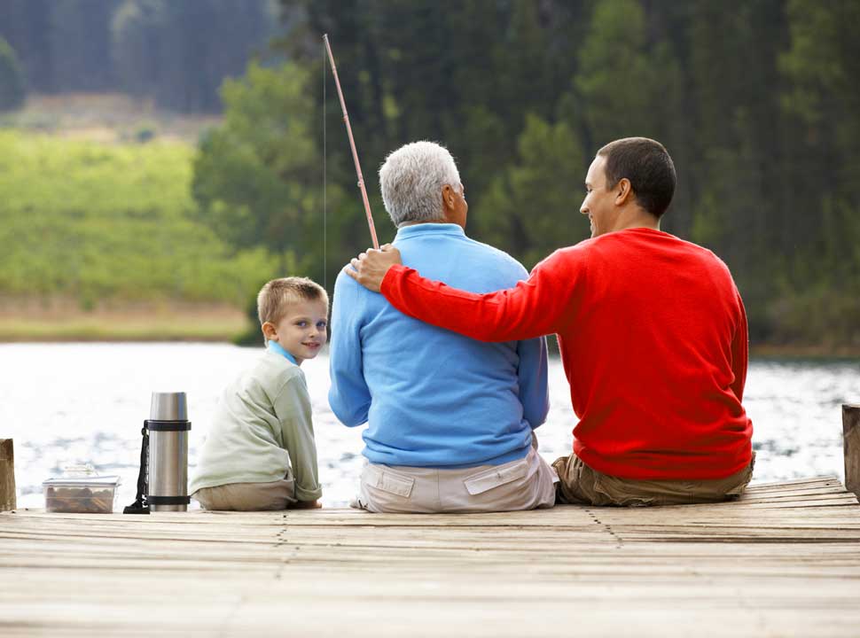 An older man sits on a pier, fishing with his son and grandson