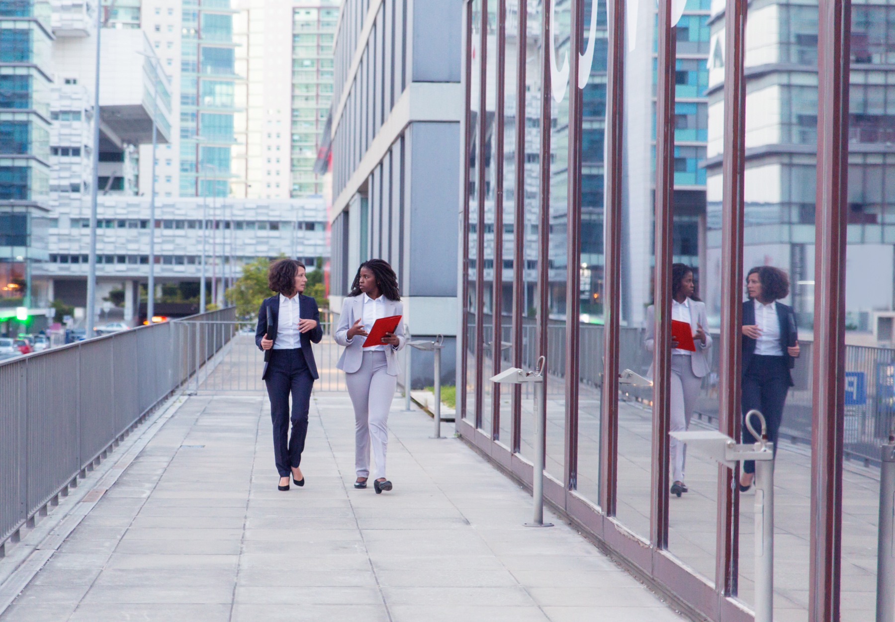 Two business professionals walking along a city office building walkway, holding documents and engaged in conversation with glass windows reflecting the scene