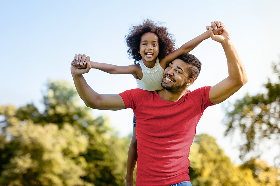 Father carrying his daughter on his shoulders while holding her hands in a green park under a clear blue sky