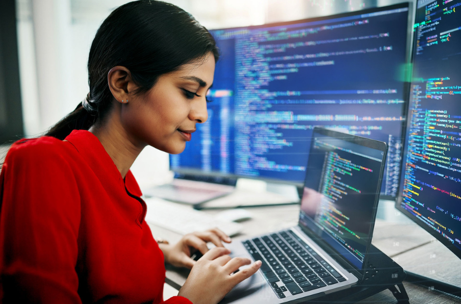 Person working on a laptop with multiple monitors displaying lines of code in a modern office setting.