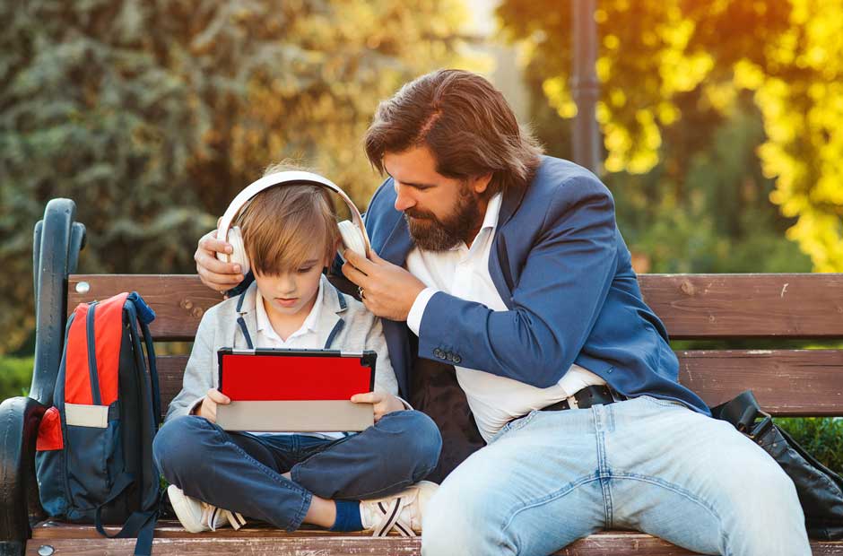 A father sits on a park bench with his son and helps the child adjust his headphones.