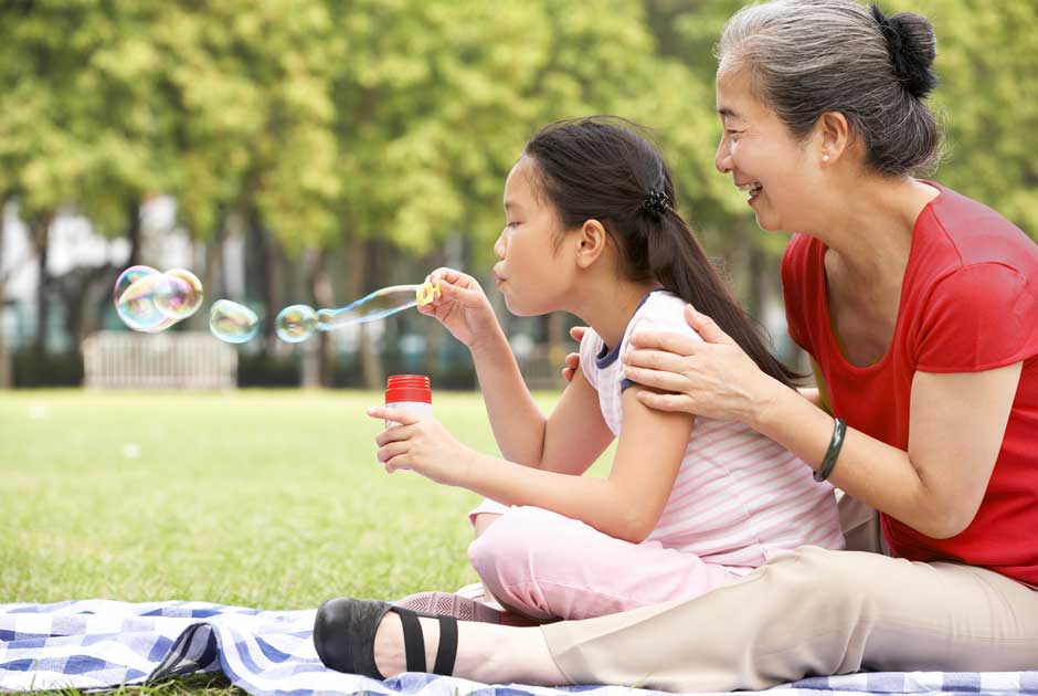 A grandmother in red and her granddaughter blow bubbles together in a park — the generations a pension promise is built to protect.