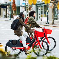 Two people ride red bicycles on a city street